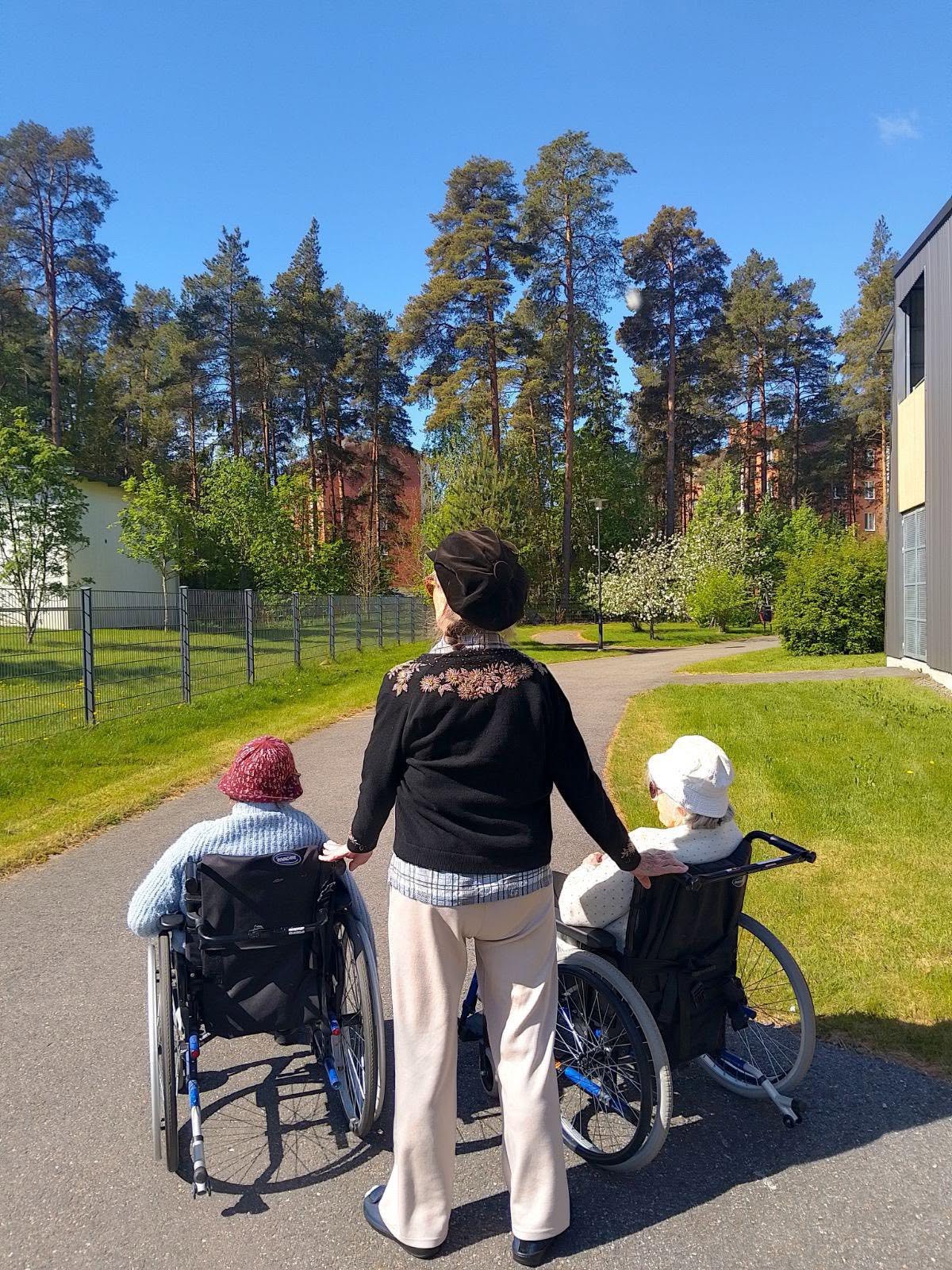 Kolme henkilöä rivissä ulkona, joista kaksi istuu pyörätuolissa ja keskimmäinen seisoo.