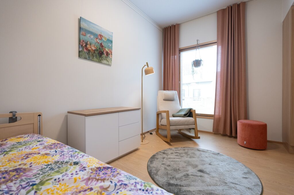 Asuinhuone, jossa on sänky, nojatuoli, lipasto ja ikkuna verhoineen.