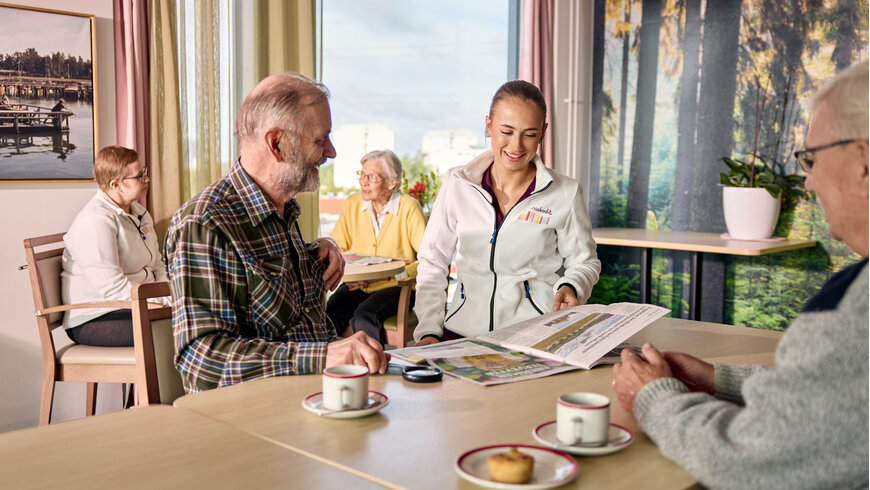 Mainiokodin hoitaja ja asukkaita kahvilla pöytien ääressä, lukevat lehteä ja juttelevat