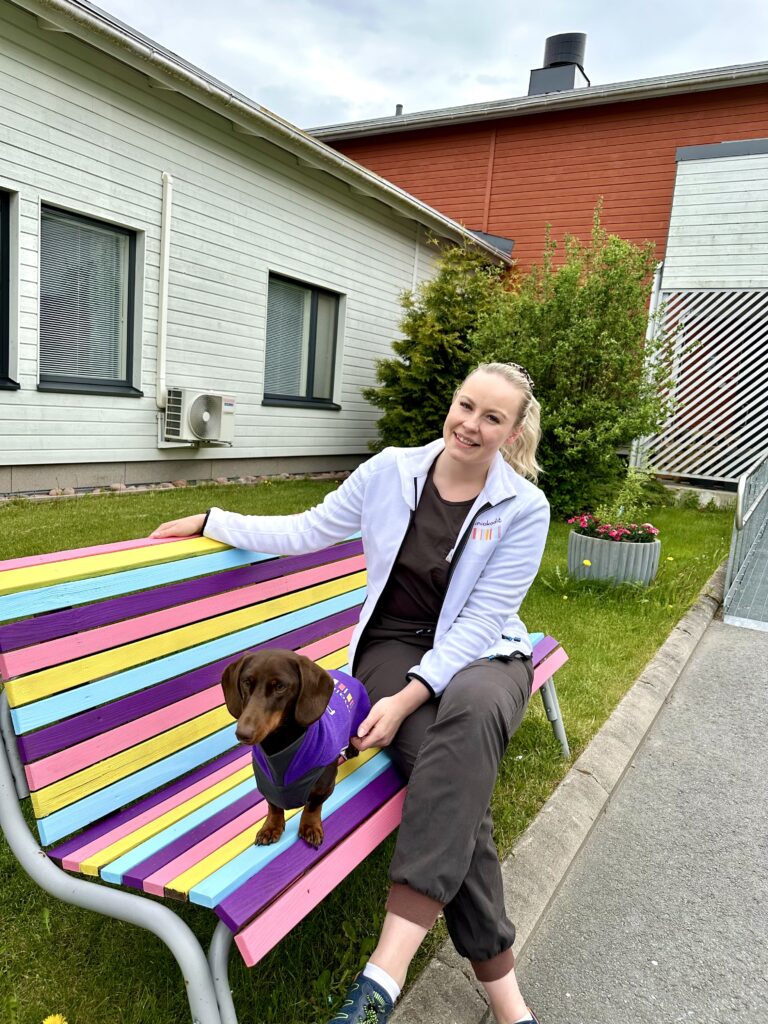 Pasi-koira ja hänen omistajansa Mikaela Jankkari istuvat värikkäällä penkillä pihalla Mainiokoti Akselin edessä. Mikaela hymyilee.